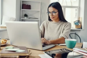 A woman with long dark hair and glasses is sitting at a desk, working on a laptop. She is surrounded by notebooks, a mug, and office supplies in a bright, modern workspace.